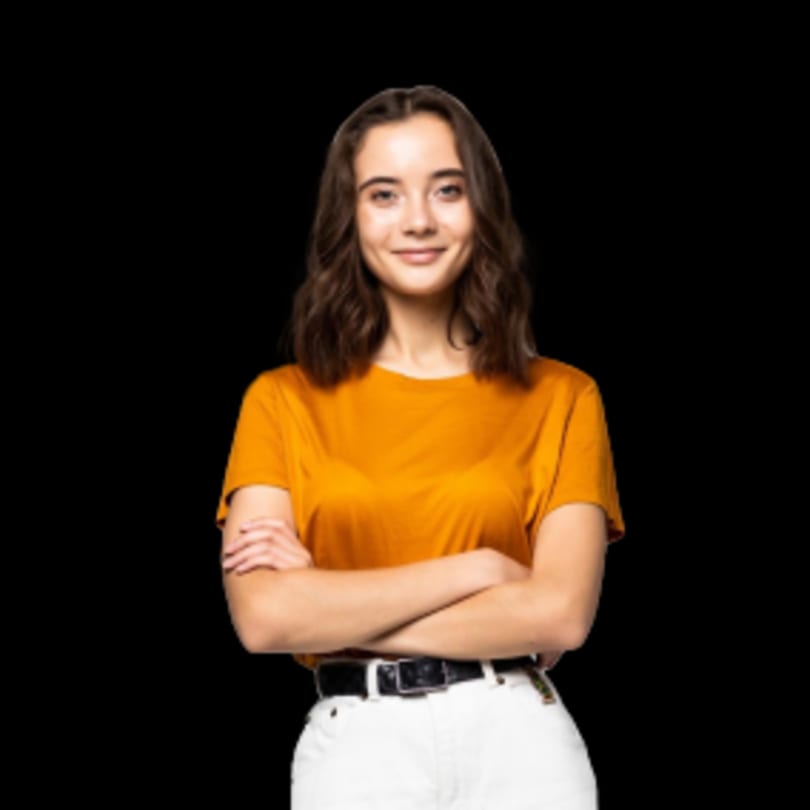 Smiling young woman with brown hair, wearing an orange shirt and white pants, arms crossed.