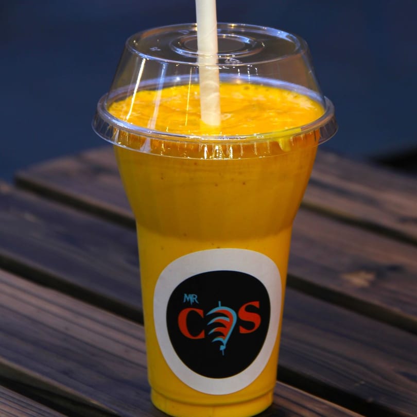 A Mr.Cone Shawarma mango smoothie in a clear cup with a straw on a wooden table.